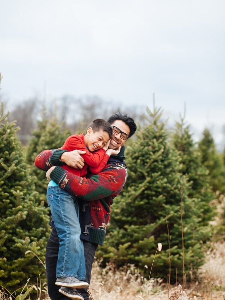 man carrying boy while standing and smiling near pine trees